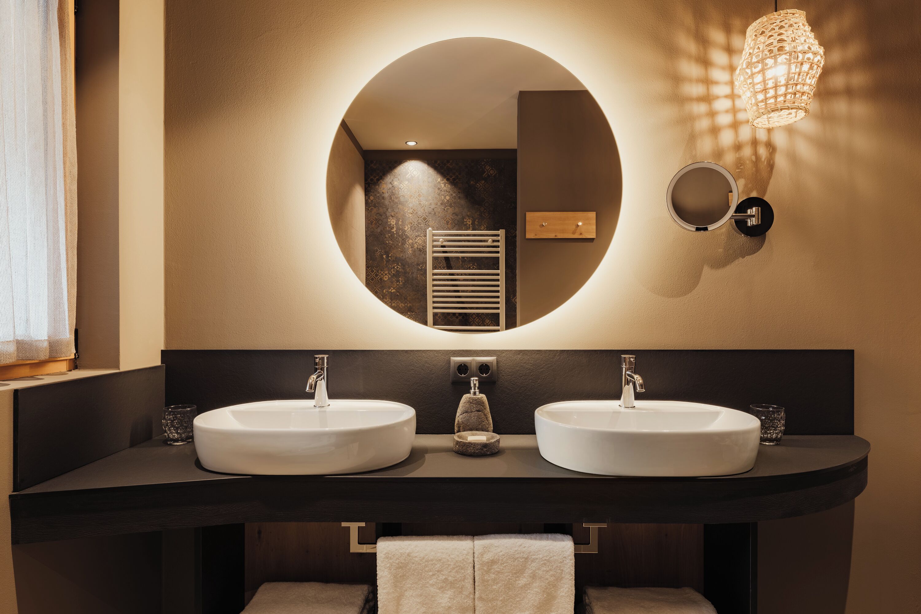 A modern bathroom with two sinks, a round mirror, and a wall-mounted light fixture.