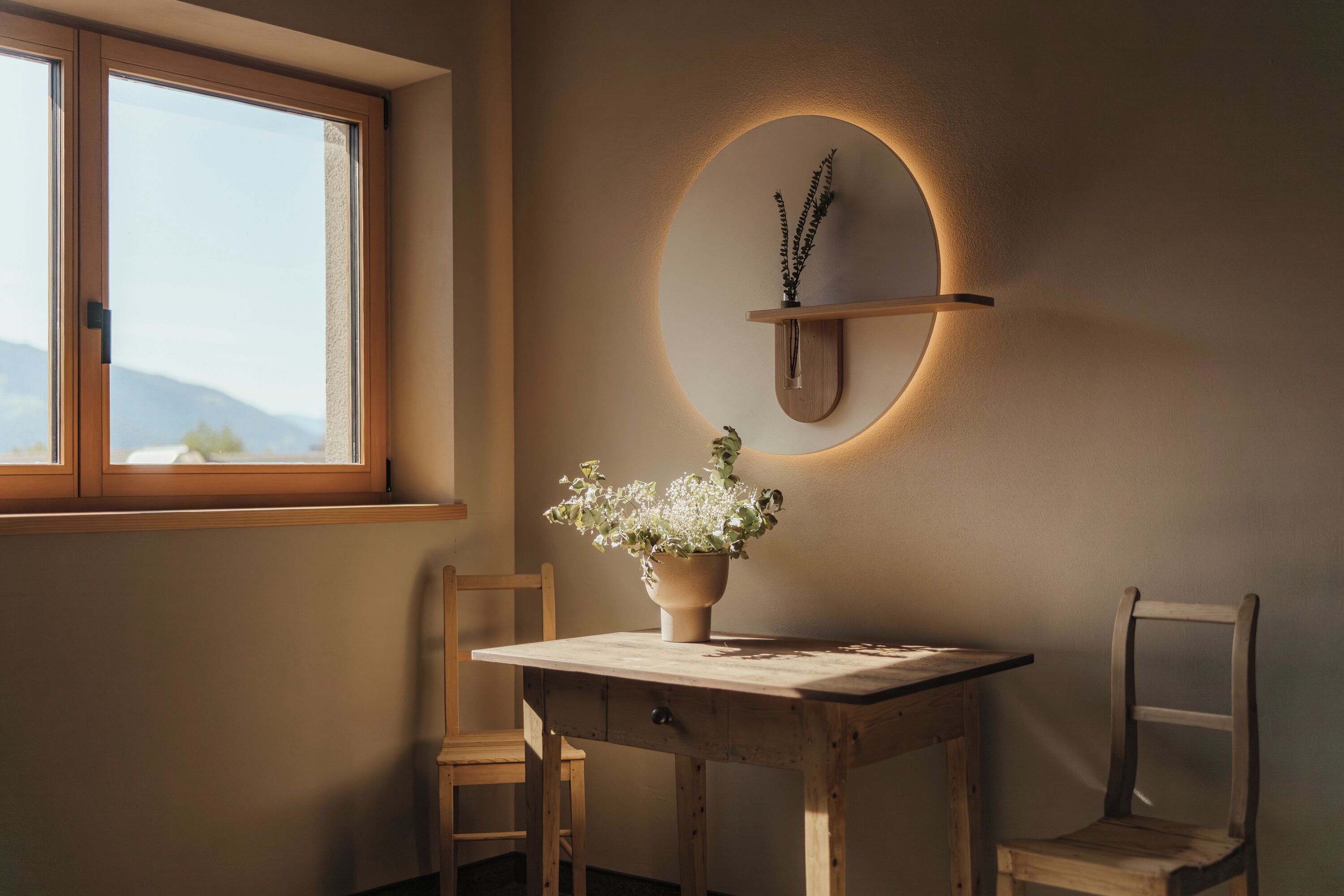 A wooden table with a potted plant, two chairs, and a wall-mounted circular light fixture.