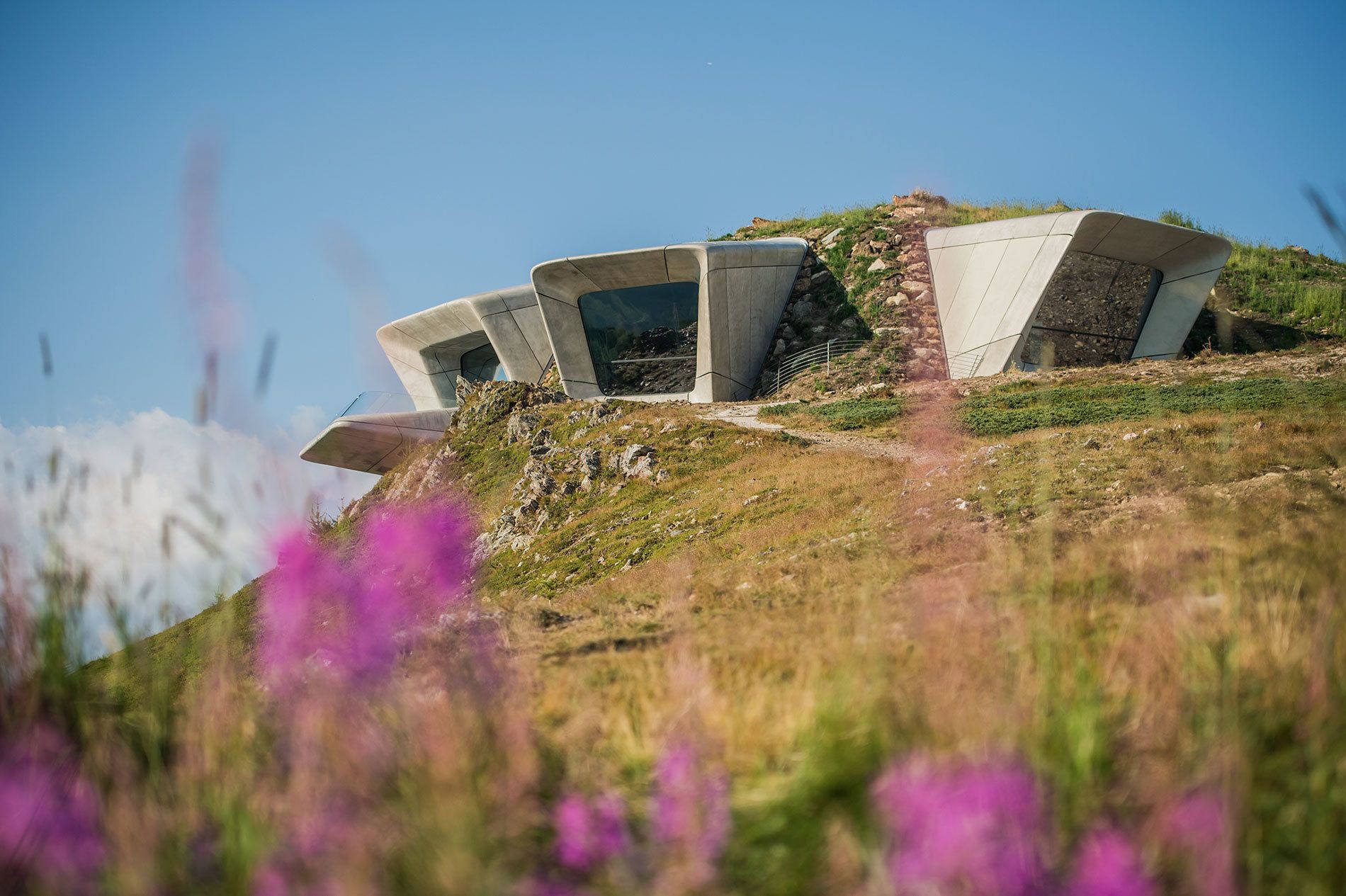A modern architectural structure with a unique, angular design, situated on a grassy hill with a clear sky in the background.