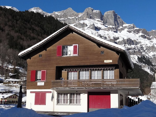 A chalet-style building with a wooden facade, red shutters, and a balcony. It is situated in a snowy mountainous area with a clear sky.