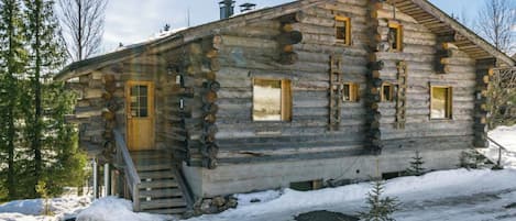 A log cabin with a wooden staircase leading to the entrance, surrounded by snow and trees.