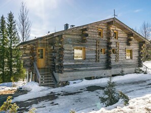 A log cabin with a wooden staircase leading to the entrance, surrounded by snow and trees.