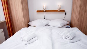 Ein ordentlich gemachtes Bett mit weißen Leinen und zwei Kissen, ein Holzregal mit zwei Wandleuchten und ein Fenster mit einem Karomuster-Vorhang.
