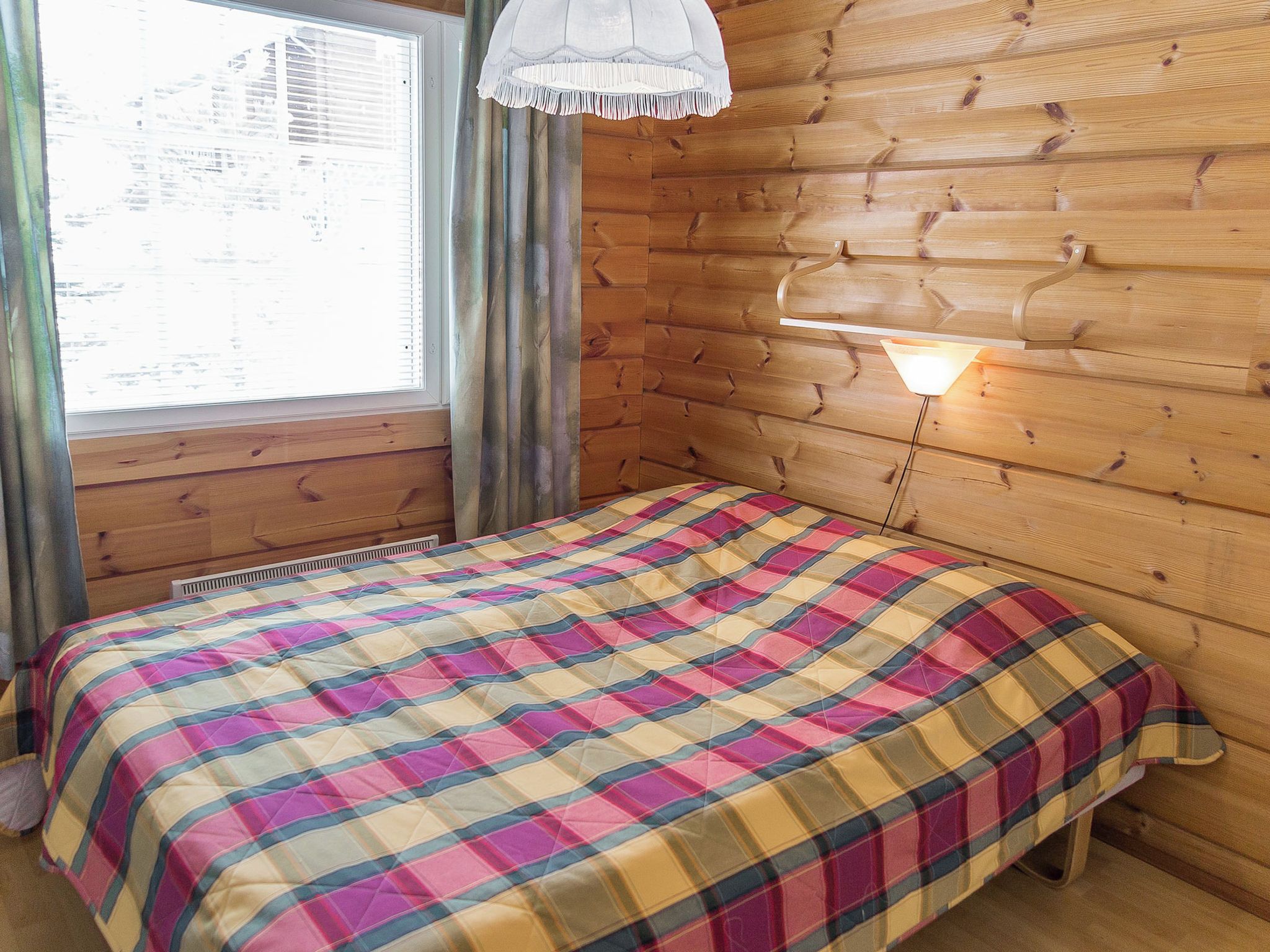 A room with a bed, a window with blinds, a wall-mounted lamp, and wooden paneling.