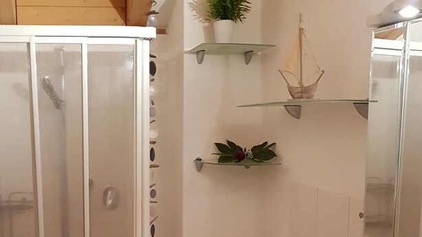 A bathroom with a glass shower enclosure, a white sink, and a wooden shelf with toiletries. - Haus Samurai by Interhome (Saas-Grund)