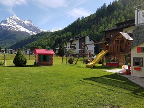 A playground with a slide, a small wooden cabin, and houses in a mountainous area.