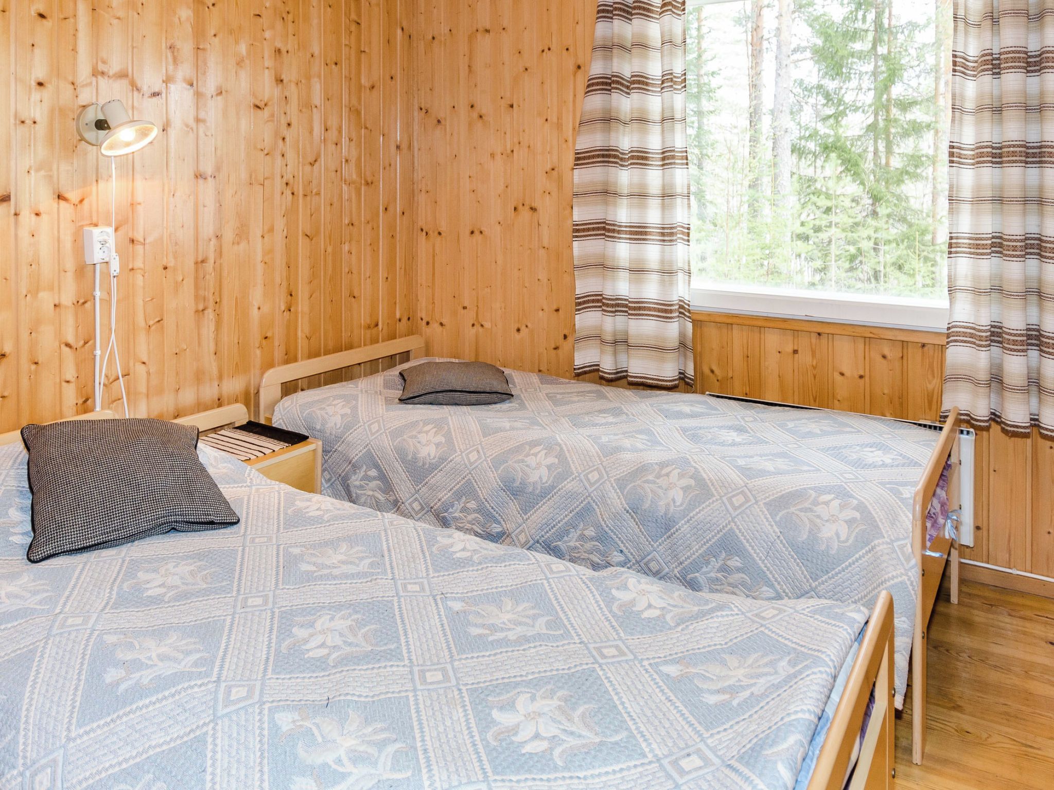 Huone, jonka seinät ovat puuta, siinä on kaksi sänkyä kuviollisilla vuodevaatteilla, yöpöytä lampun kanssa ja ikkuna, jossa on verhot.
