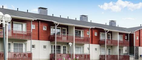 Prédios de apartamentos em tijolos vermelhos com varandas, uma entrada com brita e um céu azul claro.