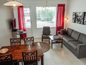 Uma sala de estar mobiliada com sofá, mesa de jantar com cadeiras, televisão e janela com cortinas.