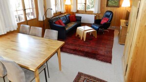 Una stanza accogliente con pavimento in legno, un tavolo da pranzo in legno, un divano e una sedia. Sono presenti finestre con tende, una lampada e un quadro appeso al muro.
