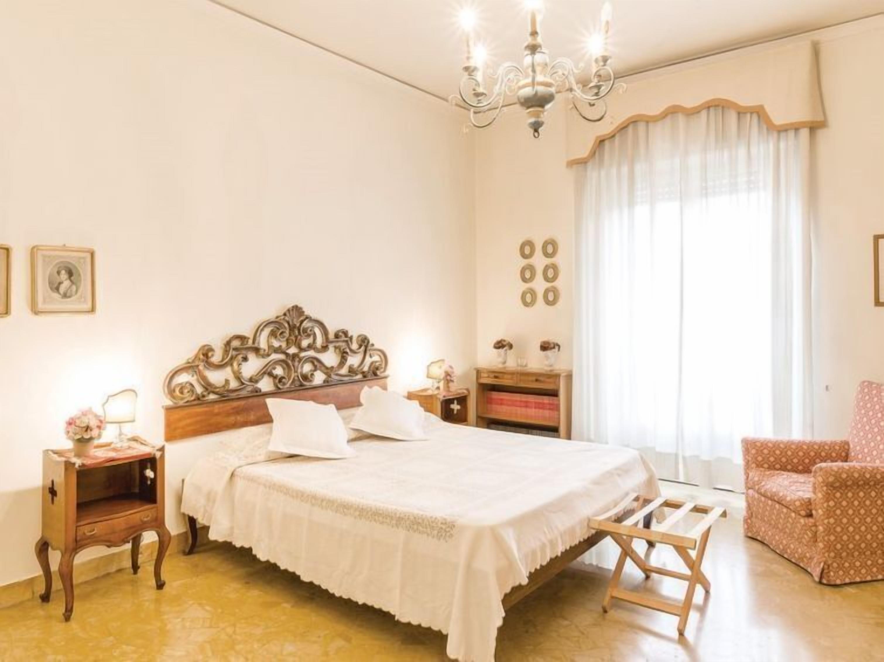 A bedroom with a large bed, a wooden nightstand, a chair, a chandelier, and a window with curtains.