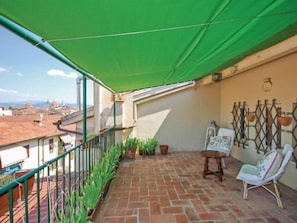 Un balcone con un tenda verde, piante in vaso, una sedia e un muro decorativo.
