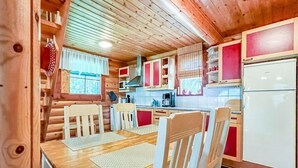 En trästuga med en matplats, bord och stolar, ett köksområde med skåp, ett kylskåp och ett fönster med gardiner.