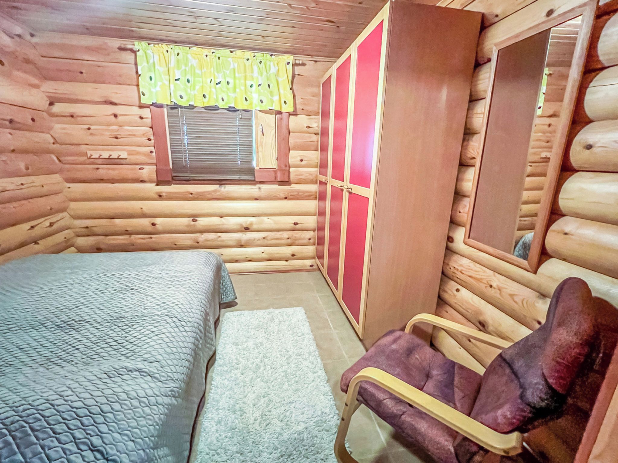 Un dormitorio tipo cabaña de madera con cama, una silla, un armario y una ventana con cortinas.