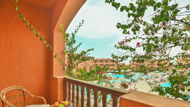Un balcone con sedie in vimini e un tavolo con frutta, vista su una piscina e degli edifici.