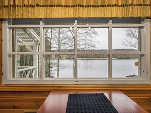 A large window with a view of a lake and trees, a wooden table in the foreground, and a yellow curtain above. - Villa siesta by Interhome (Hämeenlinna)