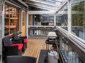 Een overdekt terras met rieten meubilair, een barbecue en uitzicht op een gebouw met grote ramen.
