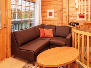 A wooden interior with a brown sofa, a round wooden table, and a window with a view of greenery.
