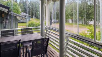 A covered outdoor seating area with a table and chairs, overlooking a forested area with a road and a car.