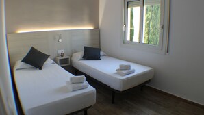 Een kamer met twee enkele bedden, elk met een kussen en een opgevouwen handdoek. Er is een nachtkastje met een lamp en een tafeltje met een decoratief object. Een raam met uitzicht op groen is zichtbaar.