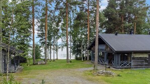 A cabin with a porch and a fenced yard, surrounded by tall trees and a body of water.