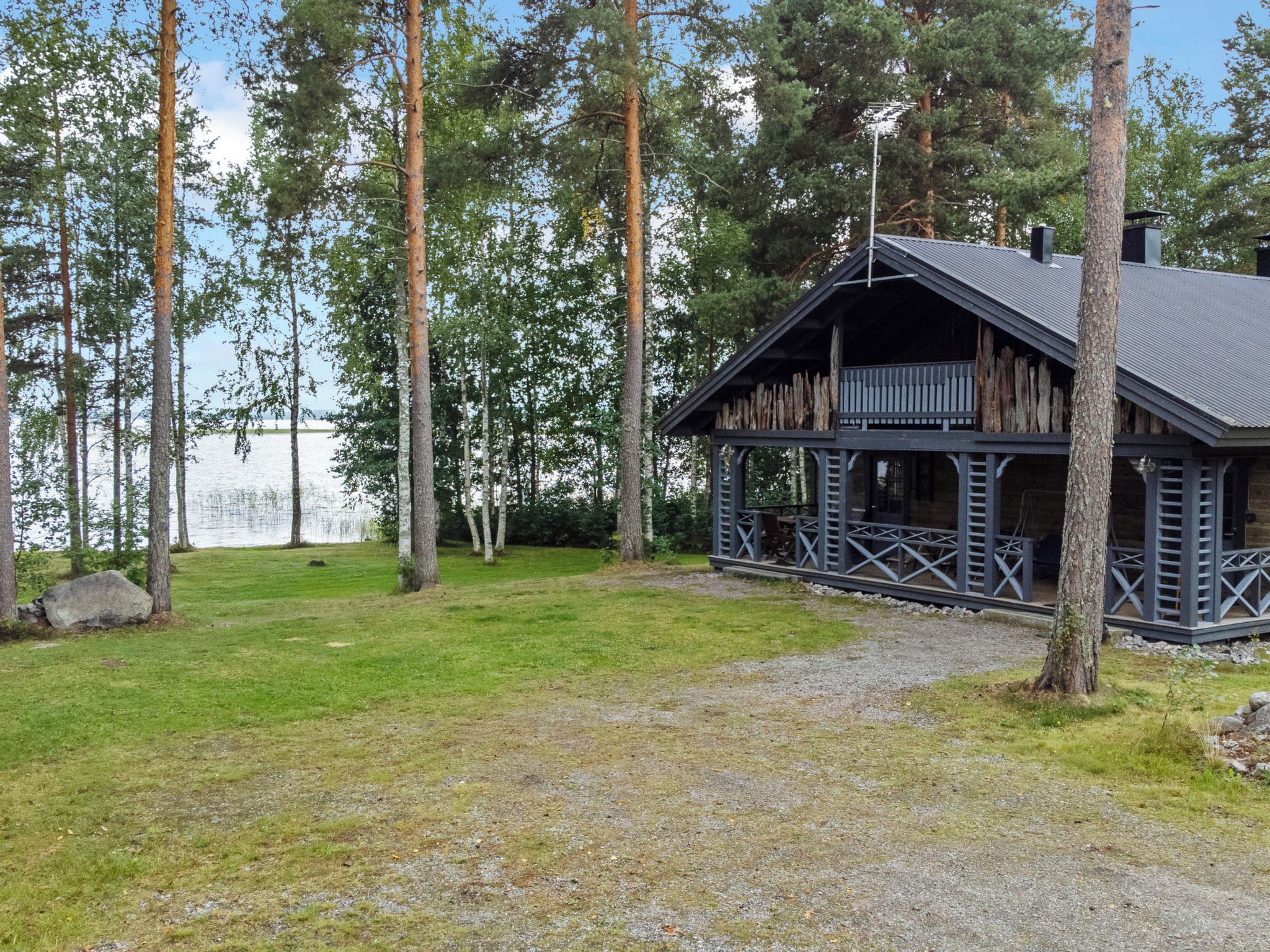 Mökki, jossa on kuisti, ja sen ympärillä on nurmikkoaluetta ja korkeita puita. Mökki sijaitsee lähellä vesistöä.