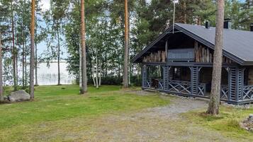 Mökki, jossa on kuisti, ja sen ympärillä on nurmikkoaluetta ja korkeita puita. Mökki sijaitsee lähellä vesistöä.