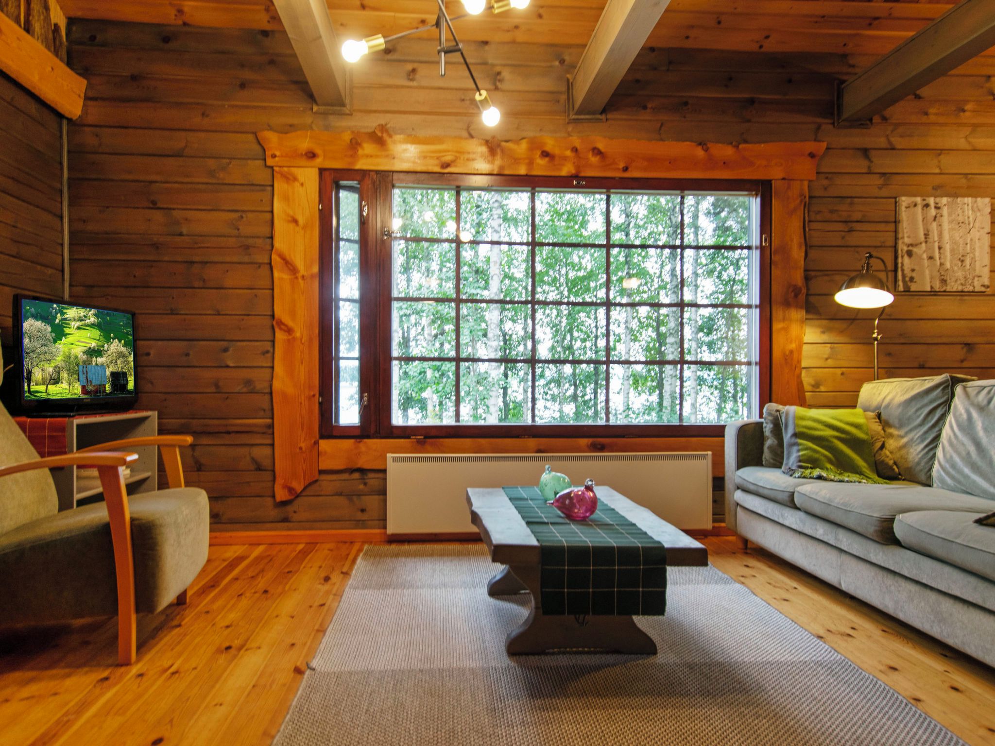 Interior acogedor de una cabaña con piso de madera, un sofá, un televisor y un ventanal con vistas a árboles.