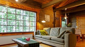 A cozy cabin interior with a wooden ceiling, a large window, a gray sofa, a small table with a green mat, and a hanging light fixture.