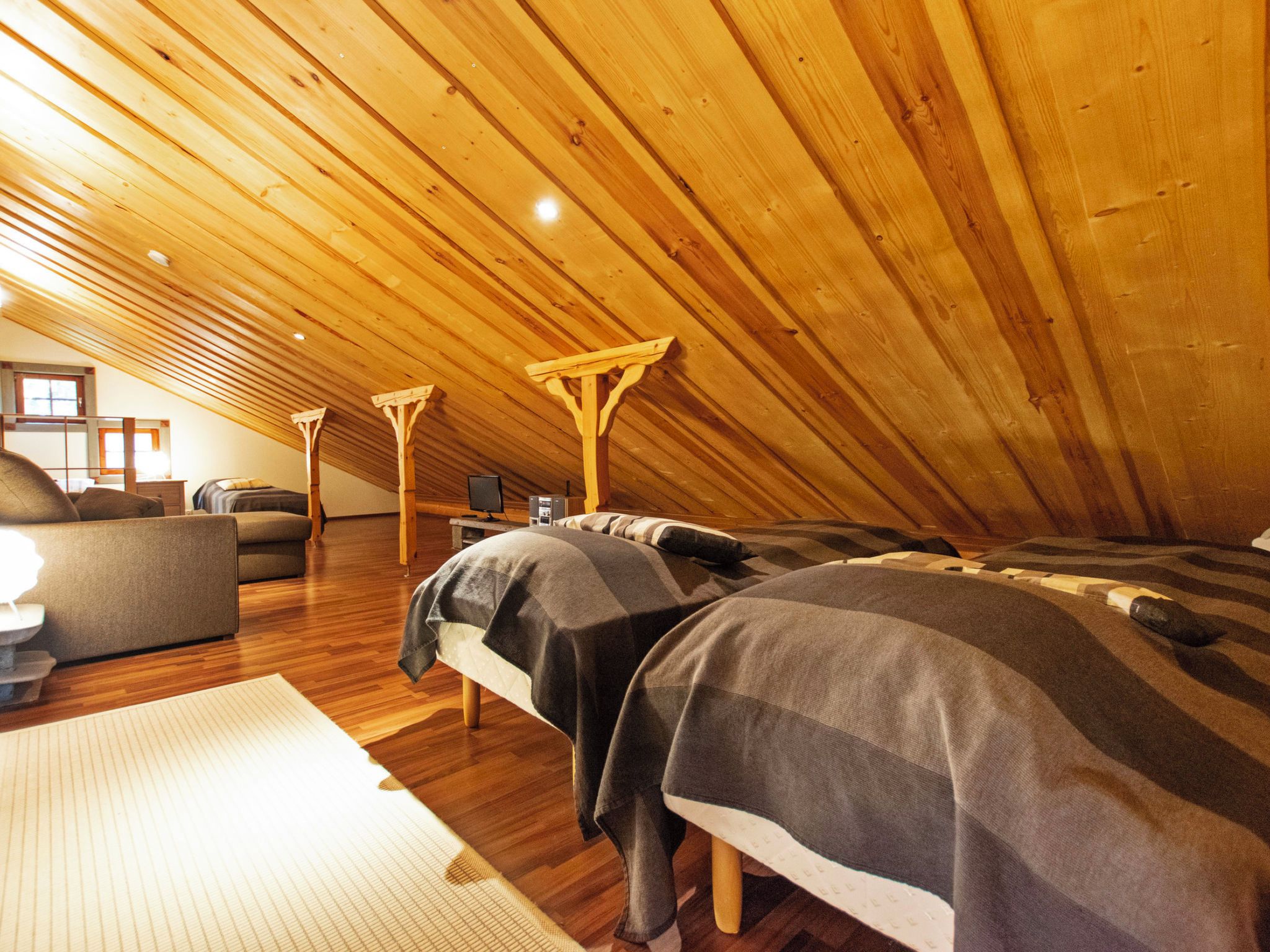 Una habitación con techos altos y una inclinación, vigas de madera, una cama con manta, una silla y una pequeña mesa.