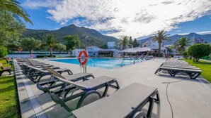 A poolside area with lounge chairs and a lifebuoy, surrounded by greenery and buildings, with mountains in the background. - Rescator Resort 115 - One Bedroom Resort, Sleeps 4 (Roses)