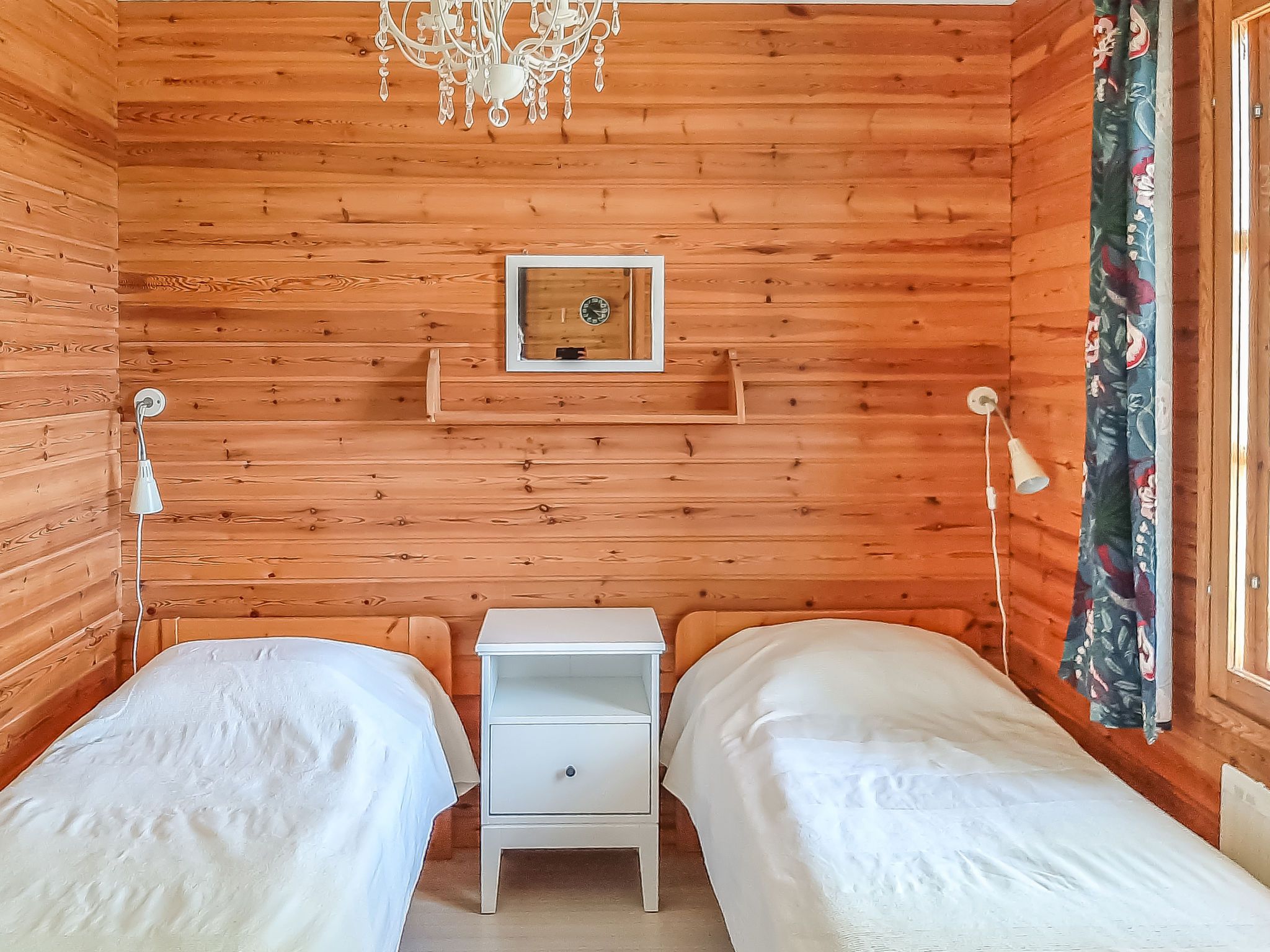 A wooden cabin room with two beds, a small white nightstand, a wall clock, and a chandelier.