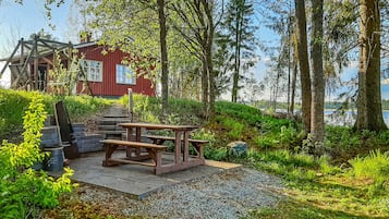 Puinen piknikpöytä penkkeineen, punainen rakennus ja metsäinen alue, josta avautuu näkymä vesistöön.