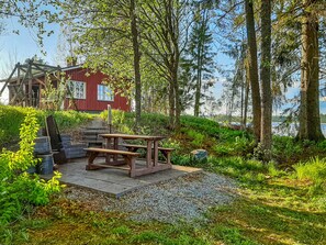 A wooden picnic table with benches, a red building, and a forested area with a view of the water.