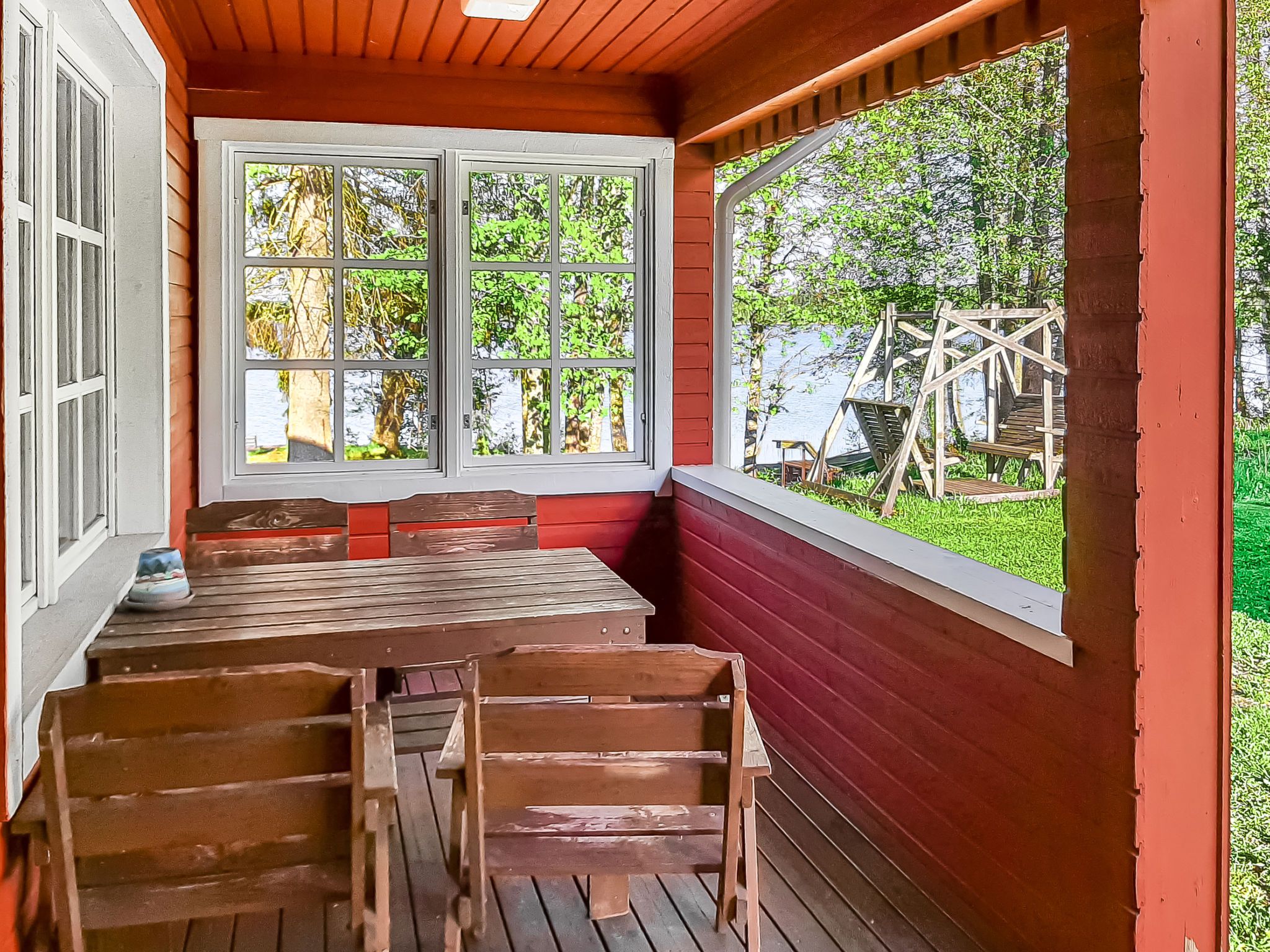 Eine überdachte Veranda mit einem Holztisch und Bänken, große Fenster mit Blick auf Grünflächen.