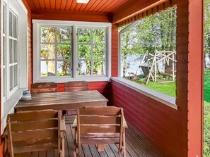 A covered porch with a wooden table and benches, large windows with a view of greenery.