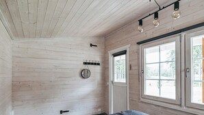 A bedroom with a wooden ceiling, a bed with a geometric patterned bedspread, a round mirror, a wall-mounted light fixture, and a window with a view of greenery. - Aapeli iso by Interhome (Ähtäri)