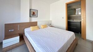A modern bedroom with a wooden headboard, a white bedspread, and two yellow pillows. There is a small bedside table with a drawer and a wall-mounted light. A framed picture hangs on the wall above the bed. - Drugstore A A - Two Bedroom Apartment, Sleeps 6 (Roses)