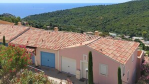 En bostadsbyggnad med ett rött tegeltak, en blå garageport och utsikt över havet och grönskande kullar i bakgrunden.