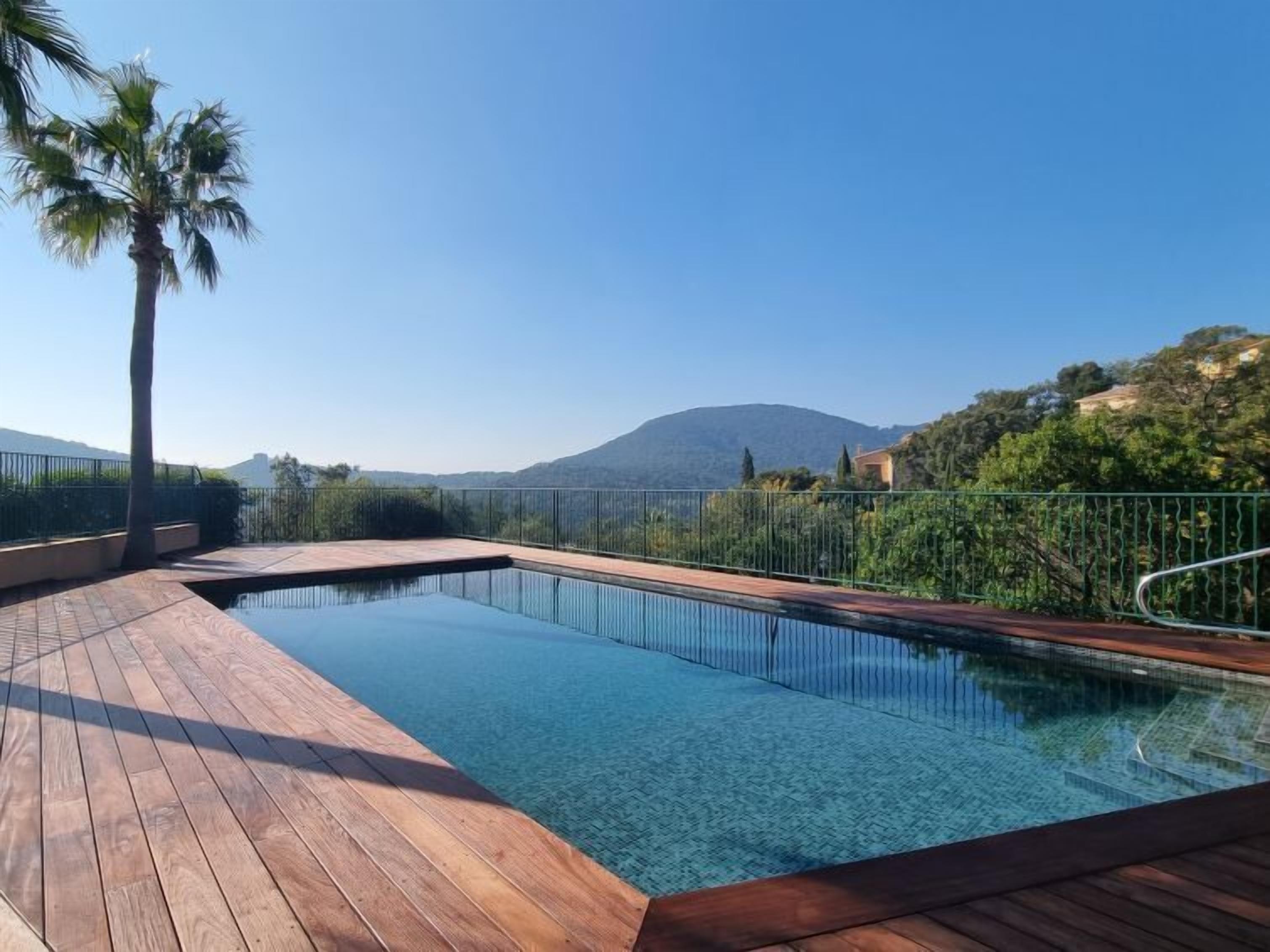 Une piscine aux eaux cristallines, dotée d’une terrasse en bois, entourée d’une clôture métallique, et offrant une vue sur une montagne et la verdure au loin.