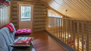 A log cabin interior with a loft bedroom, a sofa with cushions, and a view of the outdoors.