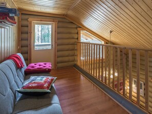 A log cabin interior with a loft bedroom, a sofa with cushions, and a view of the outdoors.