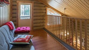 Ein gemütliches Blockhaus-Interieur mit Holzbalkendecke, einer Couch mit gestepptem Kissen, einem kleinen Fenster und einem Balkongeländer.