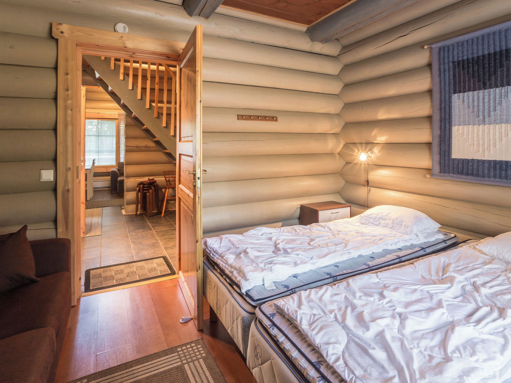 Ein Blockhauskamin mit einem Bett, einem Nachttisch, einer Wandleuchte, einer Treppe und Blick in den angrenzenden Raum.