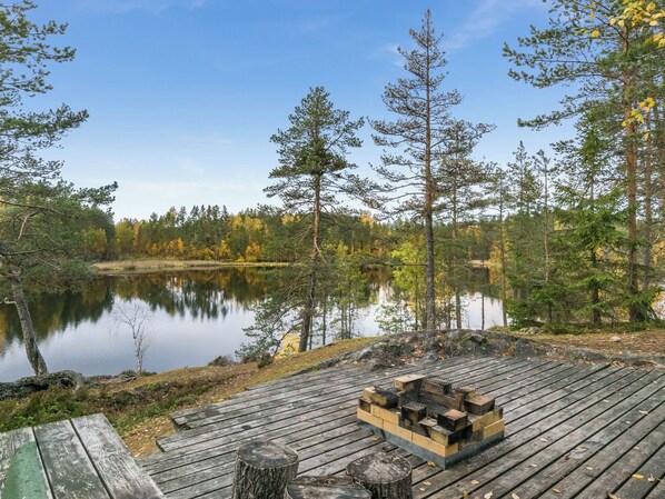 Una terraza de madera con un pozo de fuego, rodeada por un bosque y un lago tranquilo.