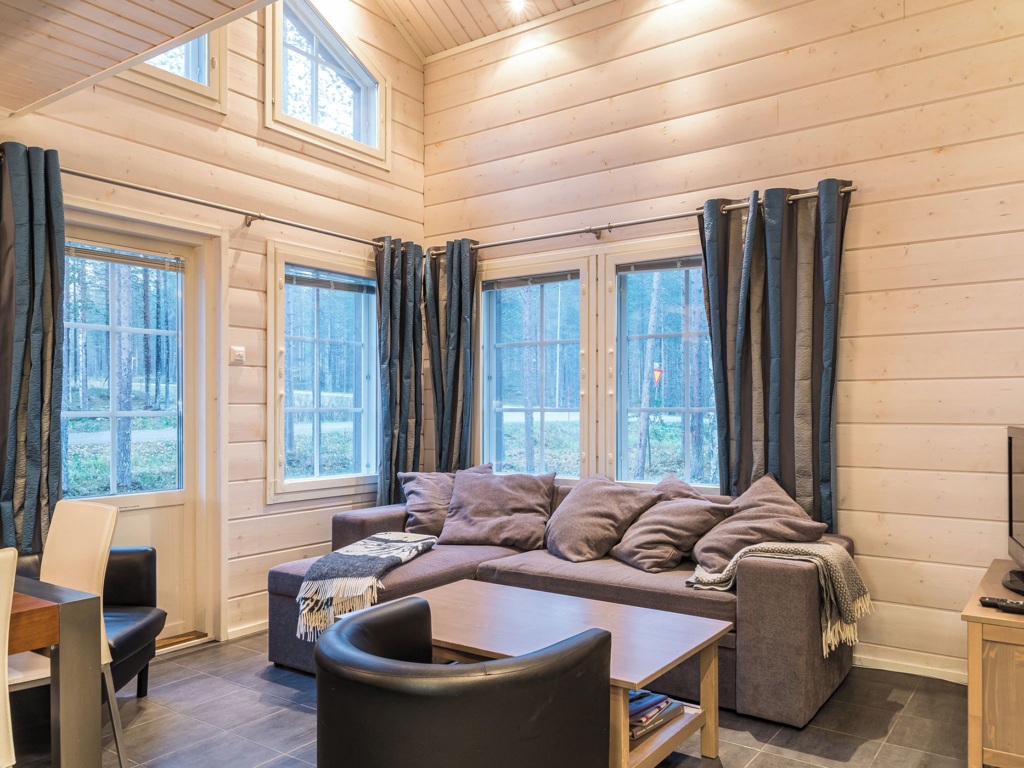 A cozy room with a sectional sofa, a coffee table, and a TV. There are large windows with curtains, and the walls are wooden.