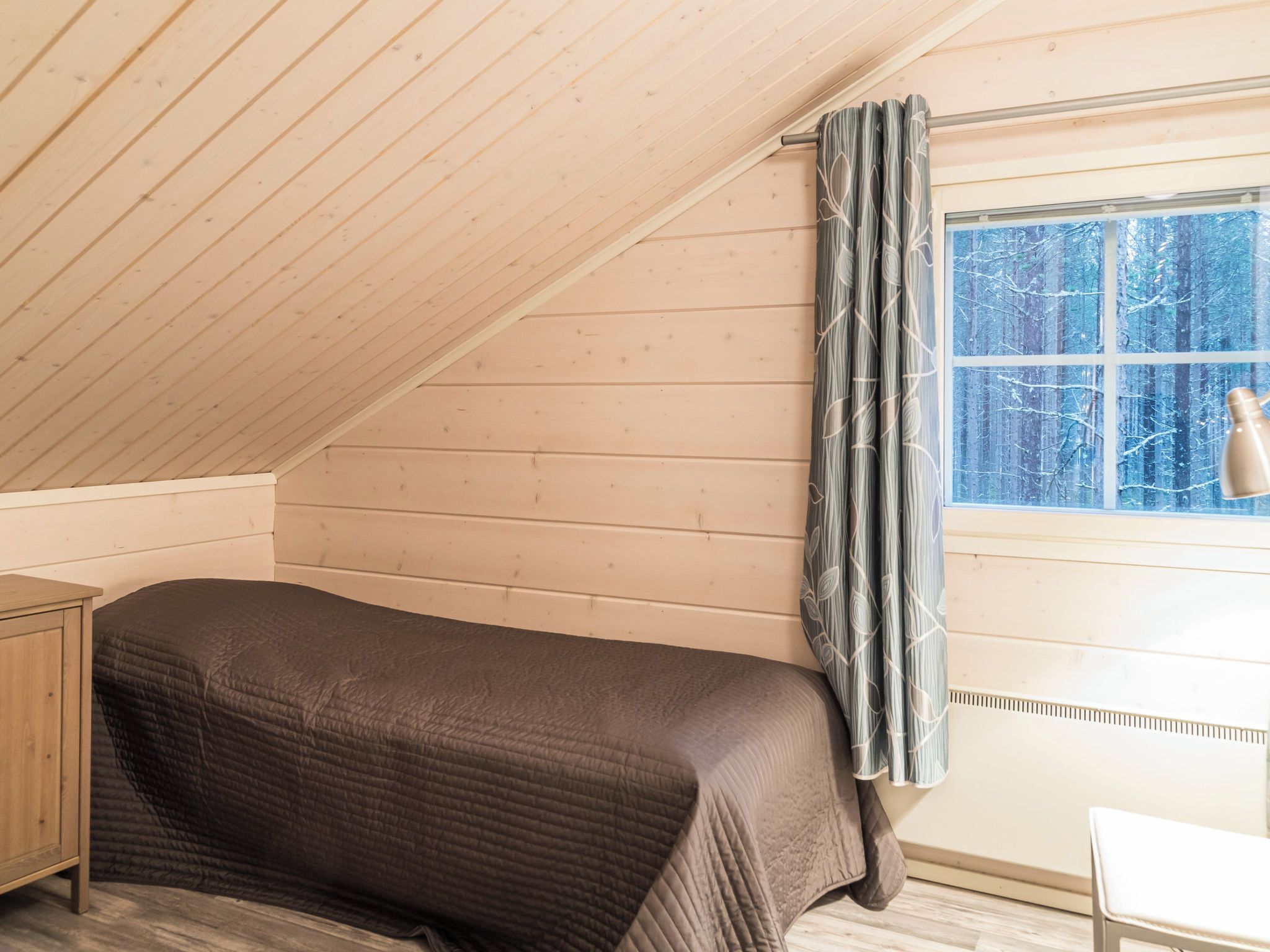 Et soveværelse med skrå vægge, en seng, et vindue med gardiner og en sengetårnslampe.