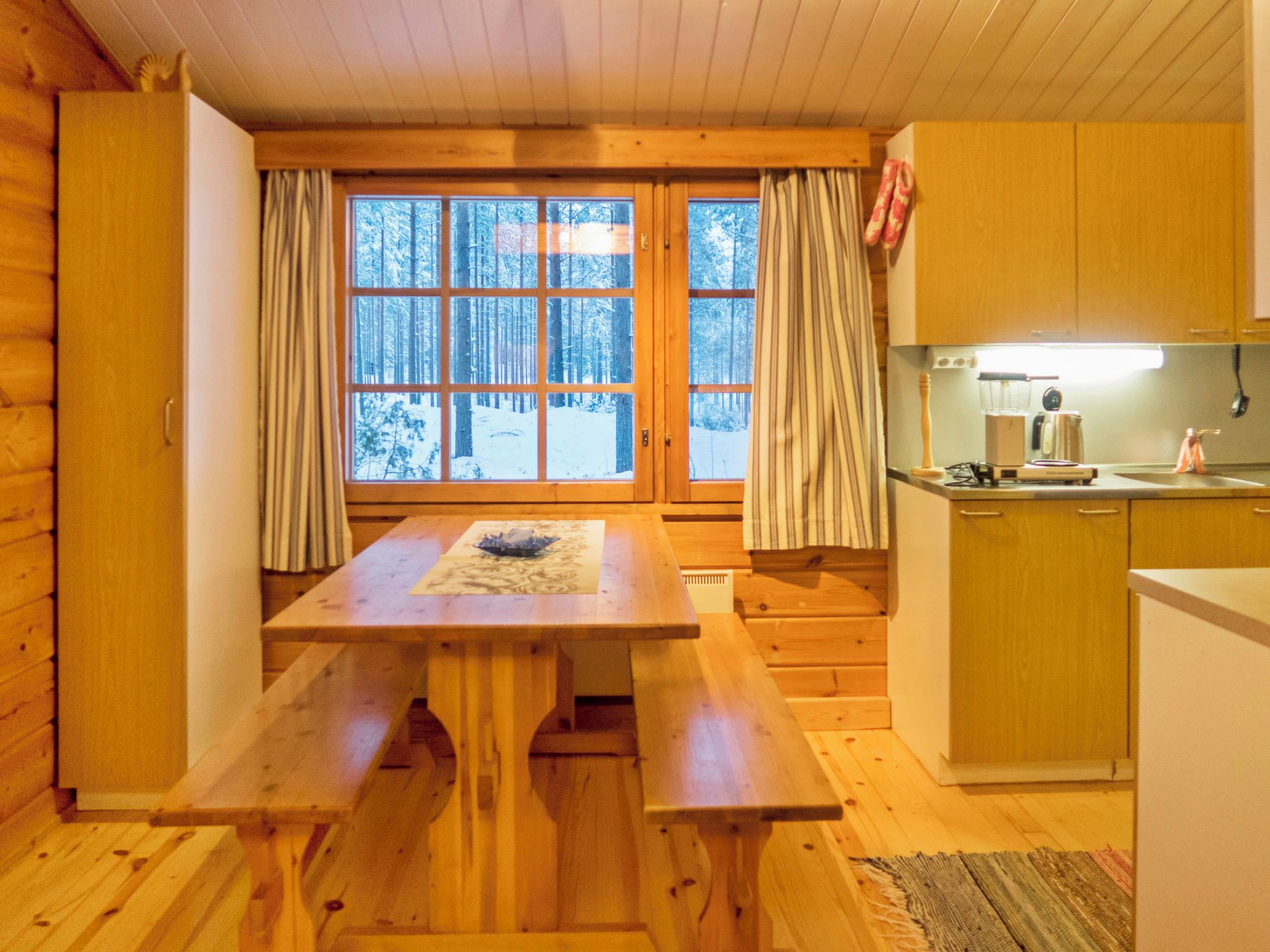A wooden cabin interior with a dining table, kitchen area, and large windows with curtains.