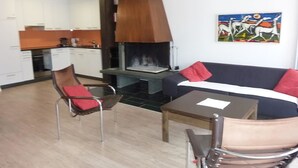 Een moderne woonkamer met een bank, een eettafel en een stoel. Achtergrond is een open haard en een keuken.