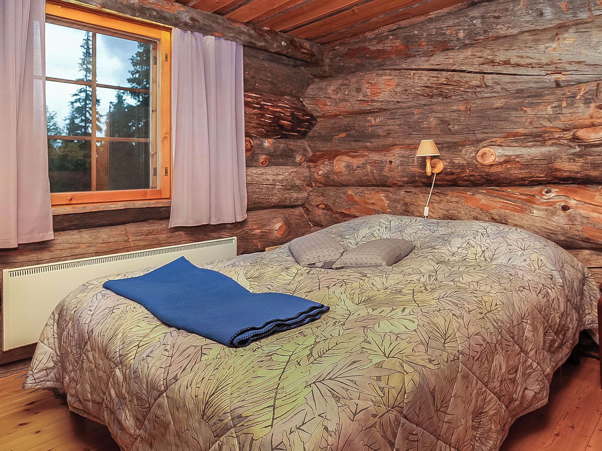 Un dormitorio de cabaña de madera con cama, ventana con cortinas y una lámpara.